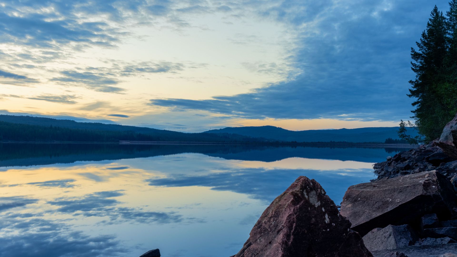 Early dawn over the Österdalälven River in Dalarna, Sweden, with calm water reflecting the blue sky and clouds. Rocks and trees frame the shoreline in the foreground, with…